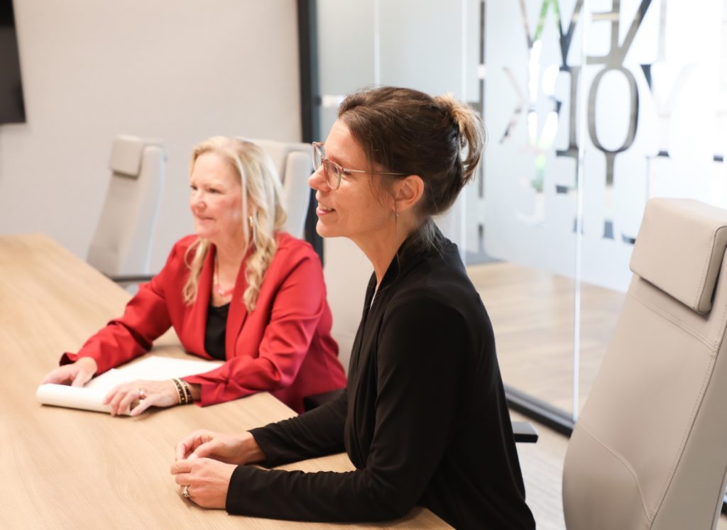 Two businesswomen in black and red blazers sitting at a conference table during a meeting in a modern office setting.