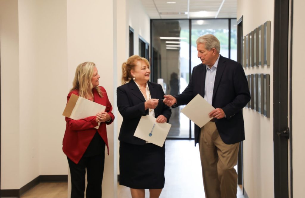 Three business professionals in formal attire walk and converse down a modern office hallway, holding folders and documents.