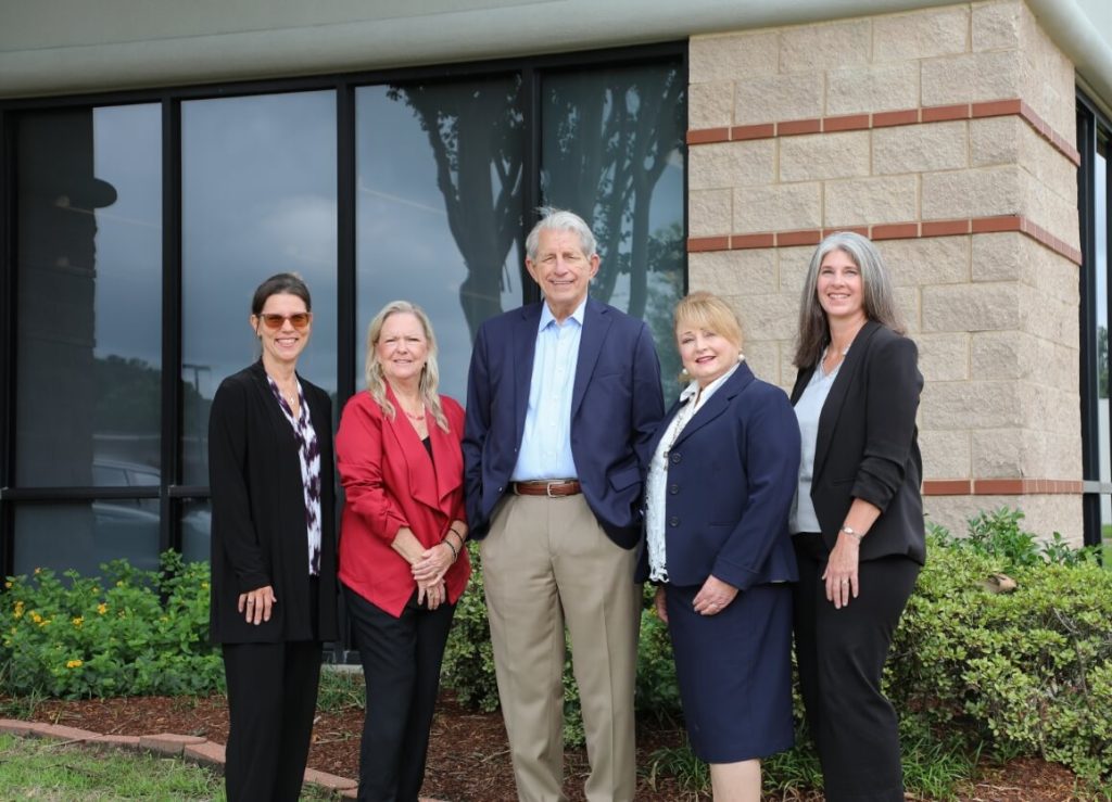 A professional group portrait of five people in business attire standing outdoors in front of an office building.