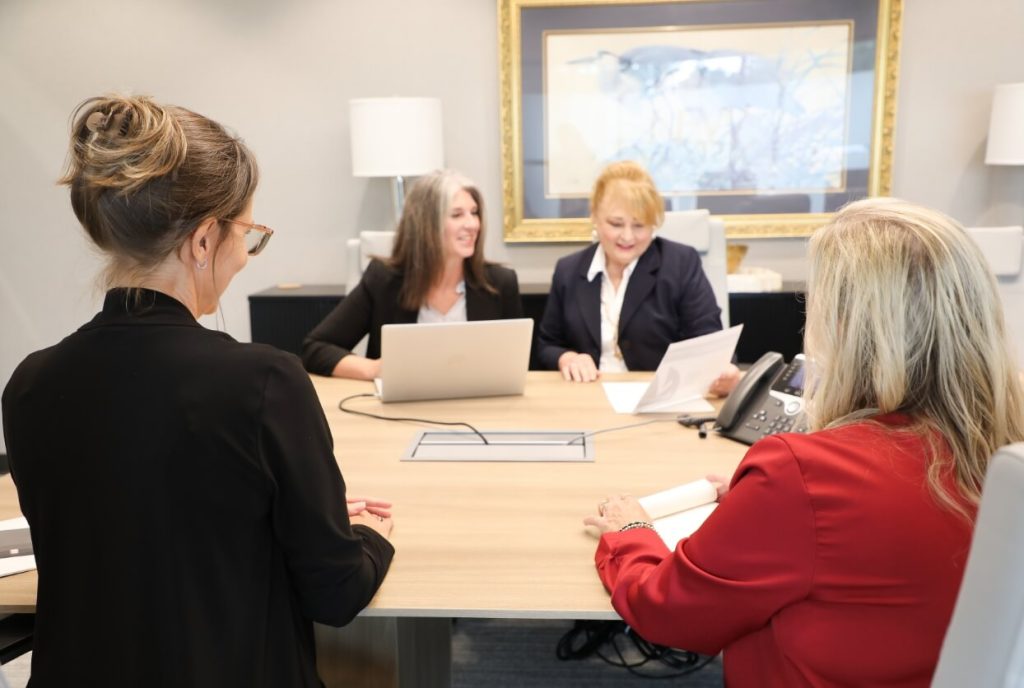 Four professional women in business attire having a positive meeting and collaborating around a conference table with a laptop and documents in an office setting.