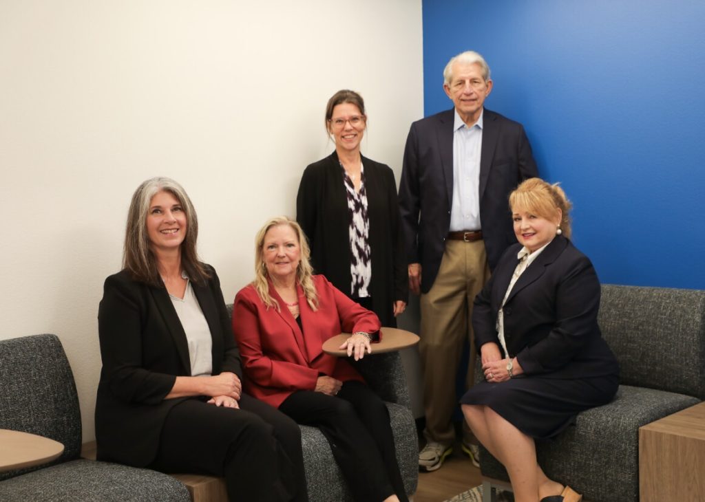 A professional group photo of the team, featuring five people in business attire posing together in a modern office setting with white and blue walls.